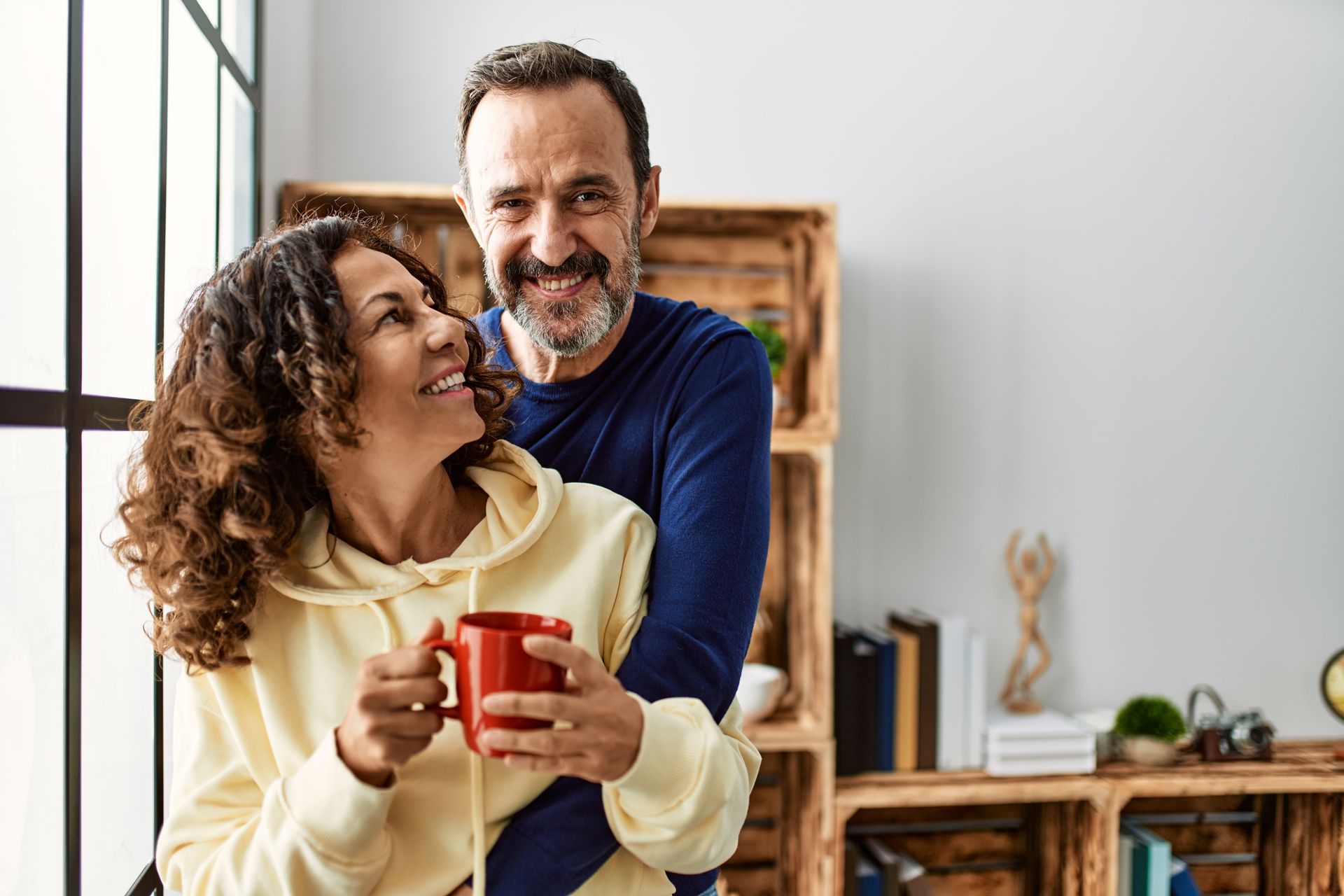 A man and a woman are standing next to each other in front of a window holding cups of coffee.