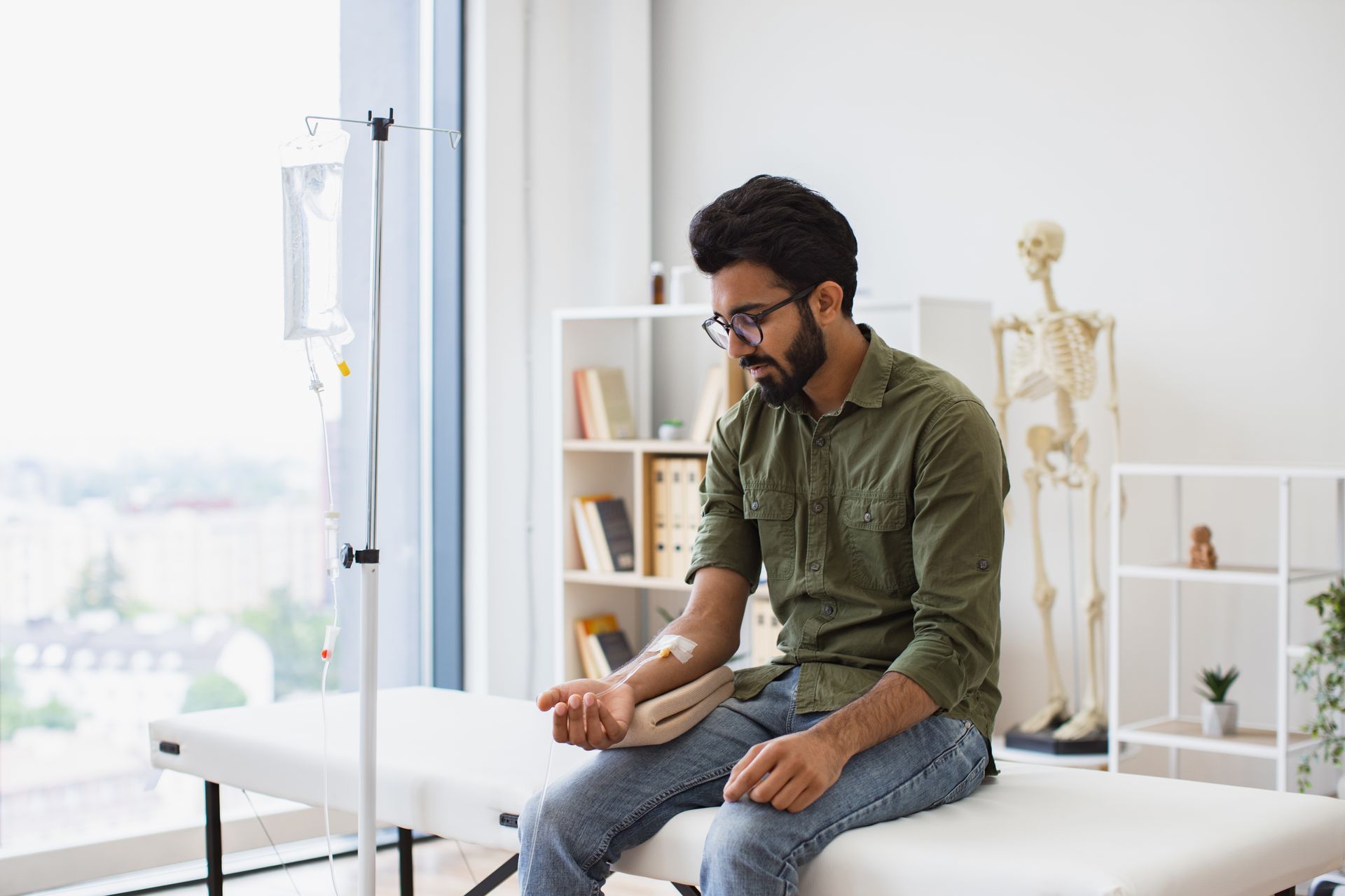 A person with glasses and a green shirt sits on a medical exam table while receiving intravenous fluids.