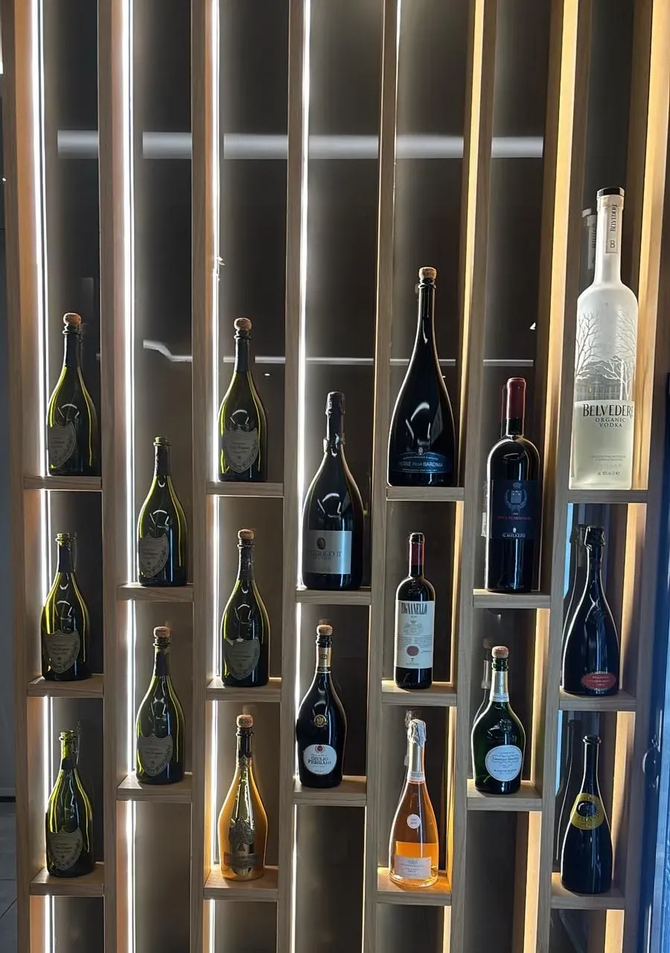 Una mensola espositiva verticale in legno con retroilluminazione, che contiene diverse bottiglie di vino e liquori in scomparti individuali.