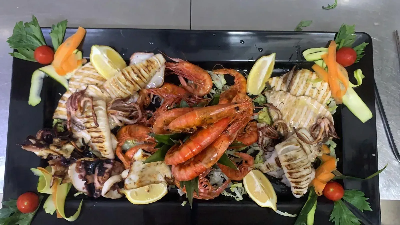Un piatto di frutti di mare grigliati, con gamberi, calamari e spicchi di limone disposti su un letto di verdure con guarnizione di ortaggi.