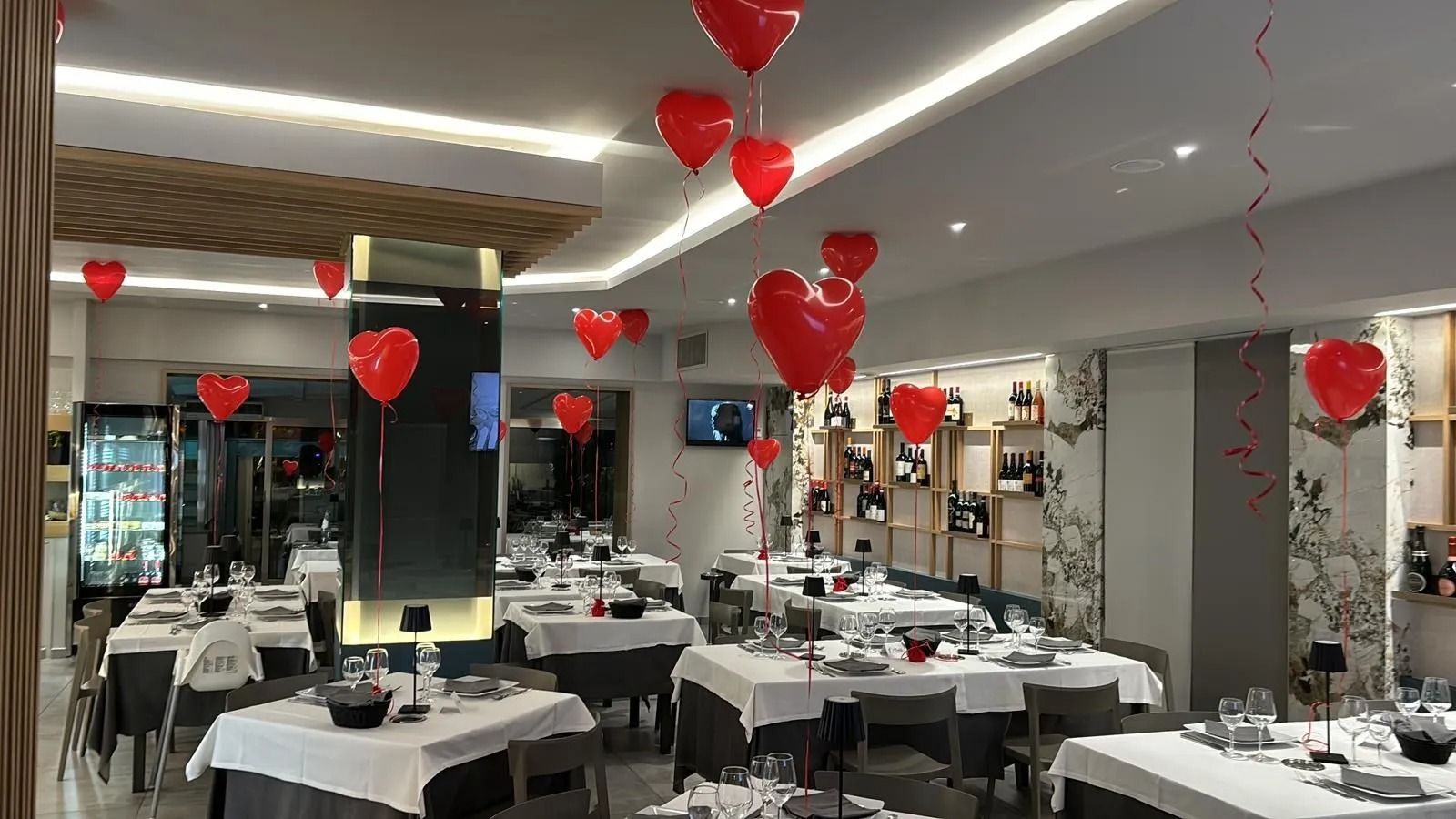 La sala da pranzo di un ristorante è decorata con numerosi palloncini rossi a forma di cuore appesi al soffitto sopra i tavoli apparecchiati.