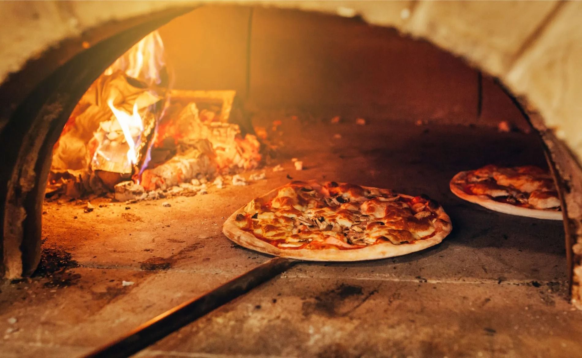 Due pizze cuociono all'interno di un rustico forno a legna in pietra, con le fiammelle che si stagliano sullo sfondo.