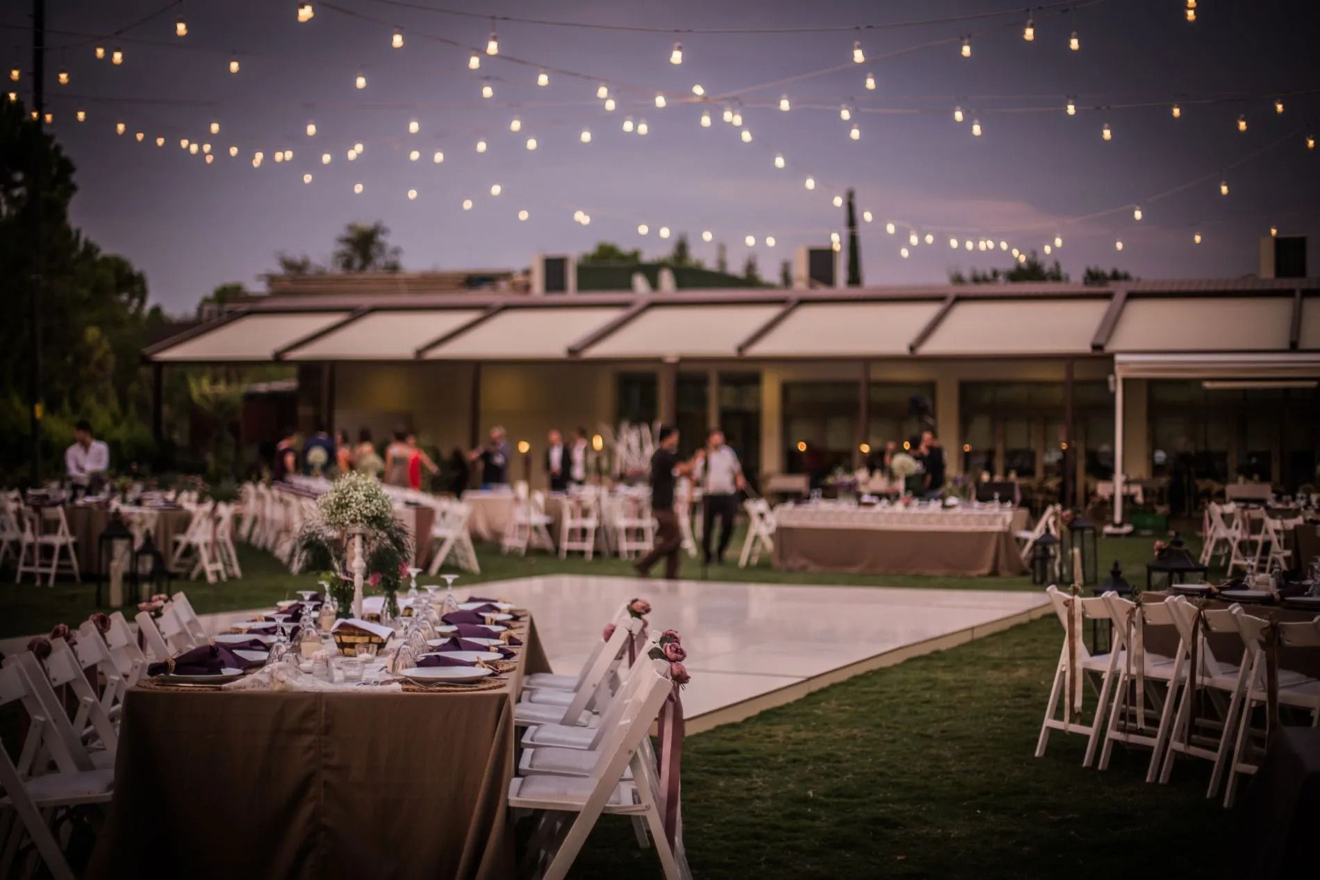 Un ricevimento di nozze all'aperto con tavoli, sedie e una pista da ballo illuminata da lucine al crepuscolo.