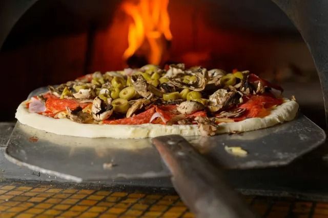 Una pizza condita con olive e funghi viene infornata in un forno a legna, con una fiamma scoppiettante sullo sfondo.