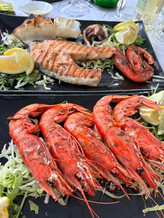 Due piatti rettangolari neri con salmone grigliato, grossi gamberi rossi e spicchi di limone su un letto di lattuga tritata.