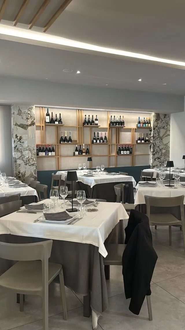 Una sala da pranzo con tavoli e sedie apparecchiati con tovaglie bianche e una mensola a muro sul fondo che espone bottiglie di vino sotto una luminosa luce a incasso.