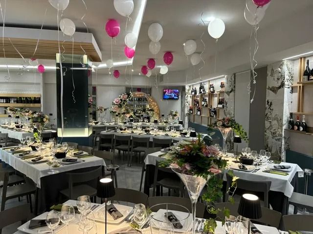 L'interno di un ristorante allestito per un banchetto con tovaglie bianche, centrotavola floreali e palloncini rosa e bianchi sul soffitto.