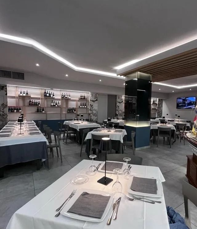 Una sala da pranzo arredata con tavoli e sedie apparecchiati con tovaglie bianche e illuminazione moderna, caratterizzata da scaffali con bottiglie di vino lungo una parete.