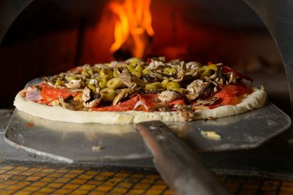 Una pizza condita con funghi e olive viene infornata in un forno a legna con una pala di metallo.