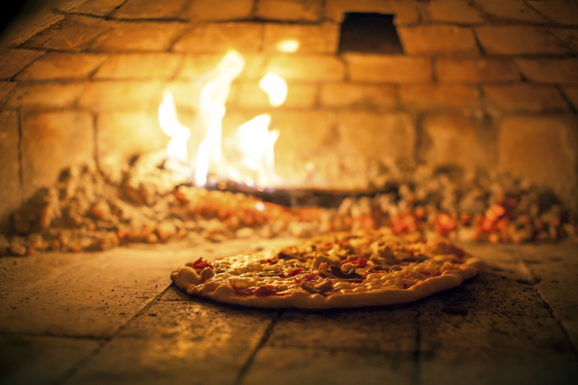 Una pizza che cuoce su un pavimento di pietra all'interno di un forno a legna, con le fiamme che brillano sullo sfondo.