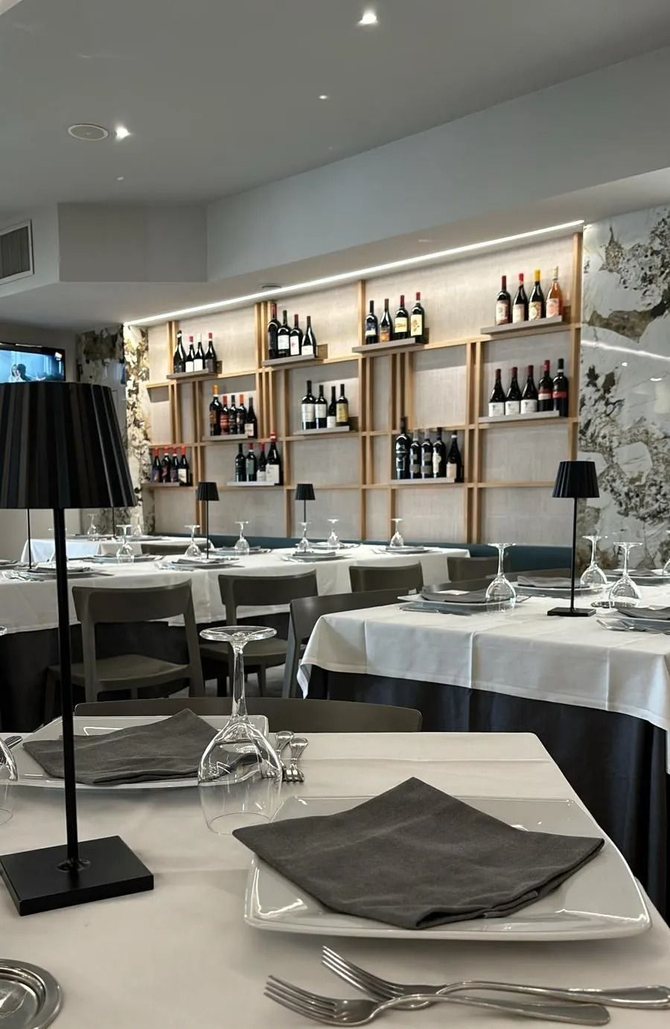 Un'elegante sala da pranzo ben illuminata, con tavoli apparecchiati con tovaglie bianche, lampade minimaliste e scaffali pieni di bottiglie di vino.