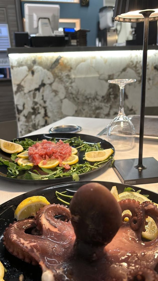 Primo piano di un piatto di polpo alla griglia, con un piatto di tartare di tonno e un bicchiere di vino sullo sfondo.