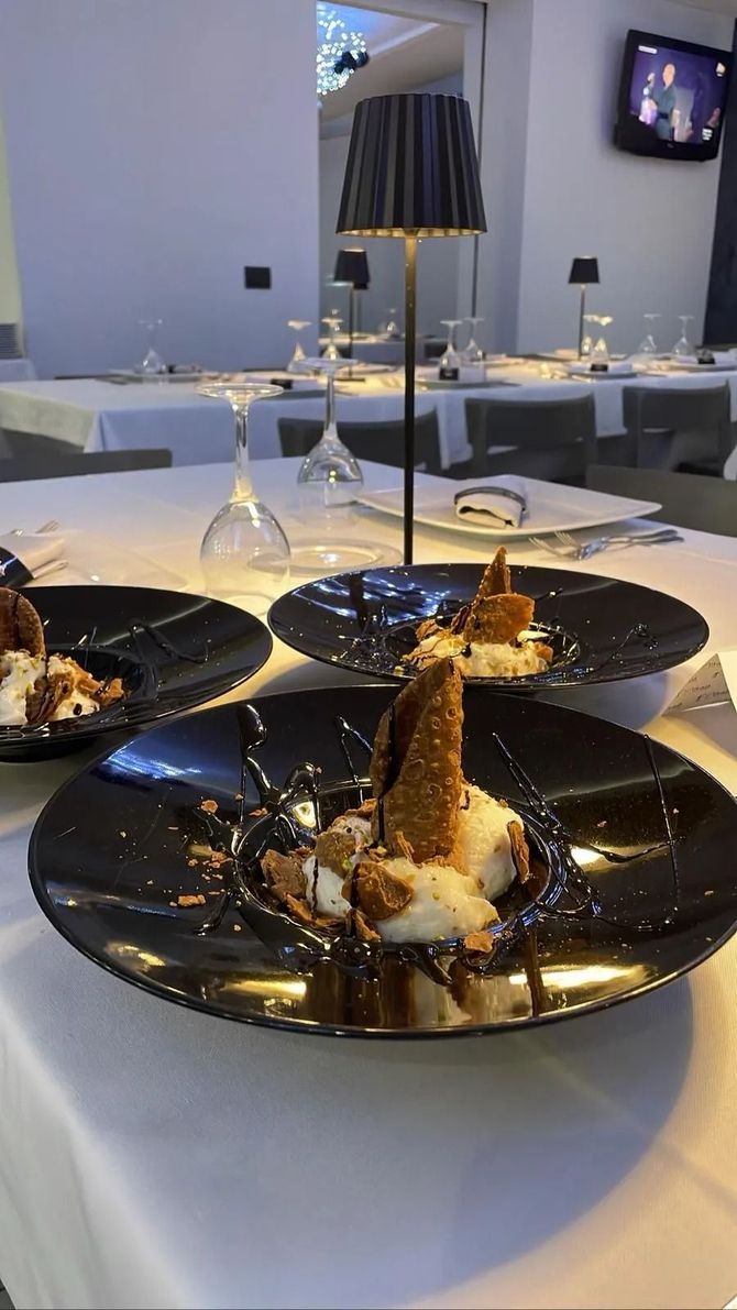 Tre piatti neri con dessert gourmet su un tavolo da ristorante apparecchiato con una tovaglia bianca, con una lampada accesa al centro.