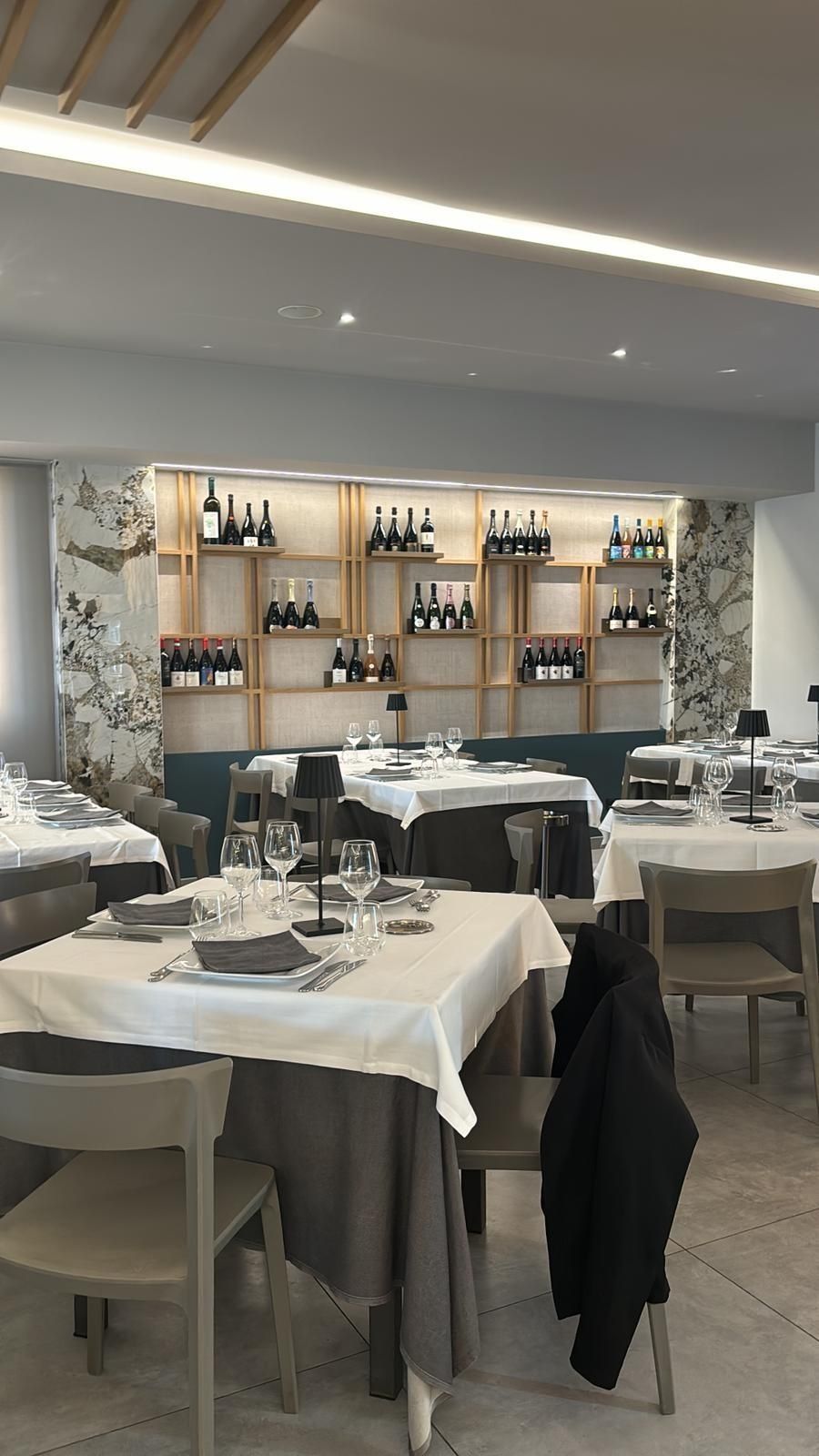 Una sala da pranzo con tavoli quadrati, tovaglie bianche e sedie chiare, e una vetrina per i vini sulla parete di fondo.