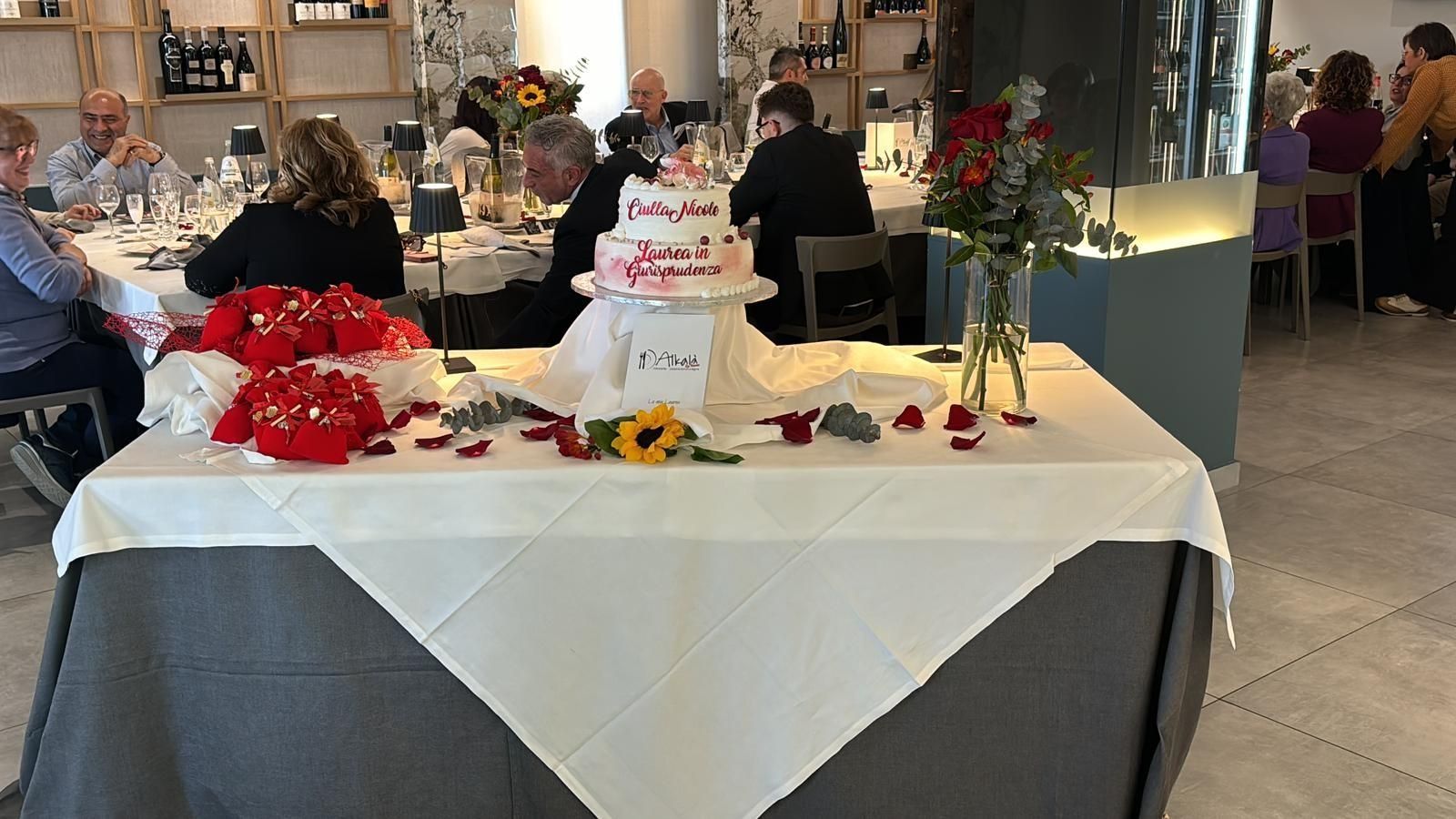 Una torta a più piani con una decorazione rossa in cima è adagiata su un tavolo drappeggiato con una tovaglia bianca, circondata da decorazioni e fiori rossi.