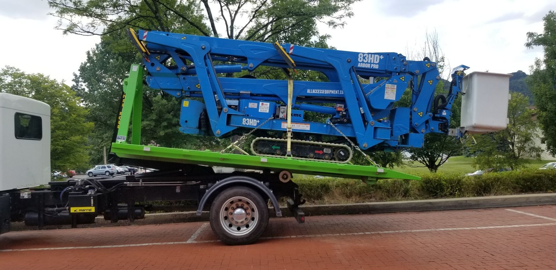 A blue aerial lift loaded on a green trailer attached to a white truck, parked on pavement.