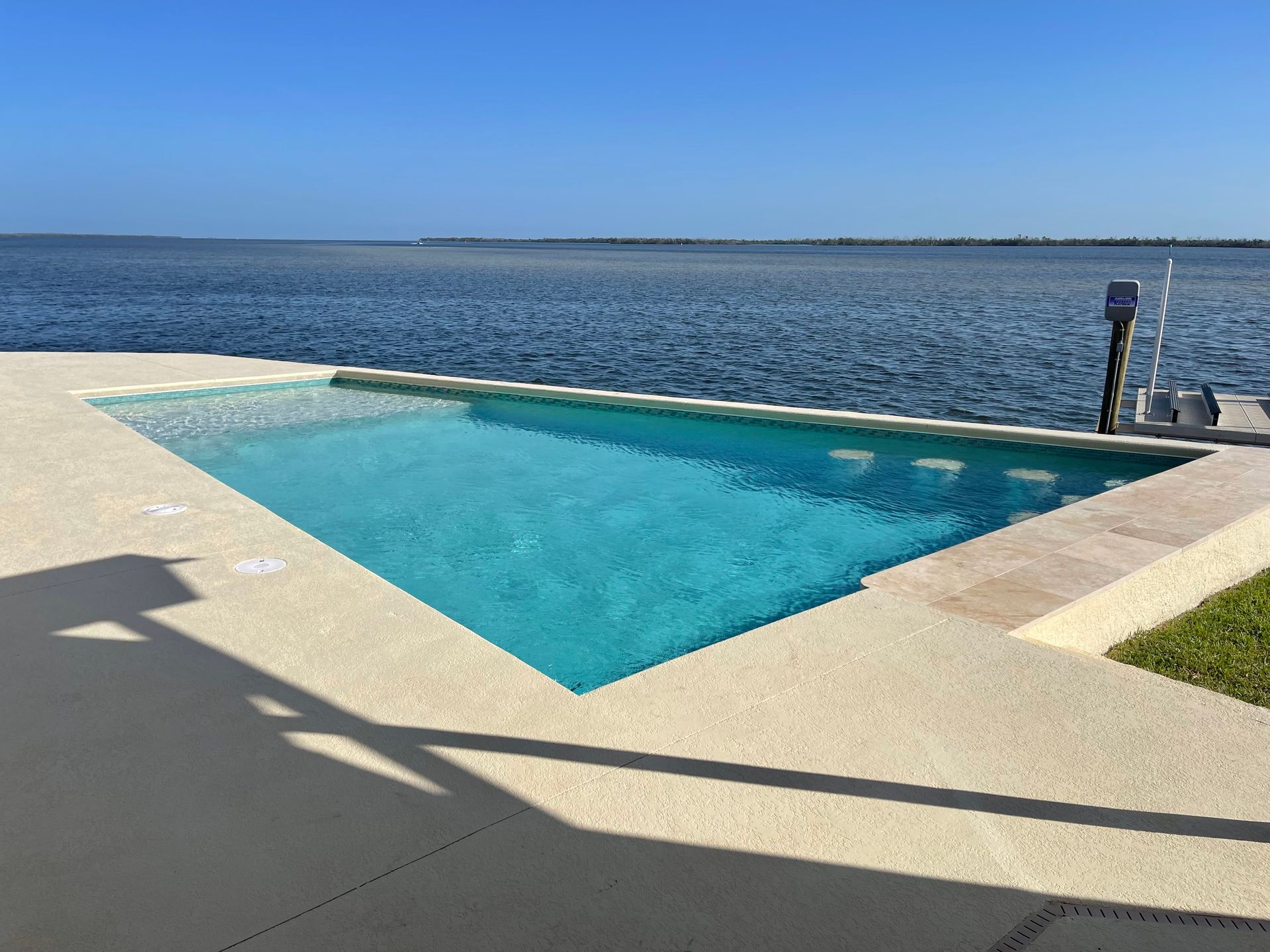 A modern, turquoise-colored pool at the edge of a large body of water under a clear blue sky.