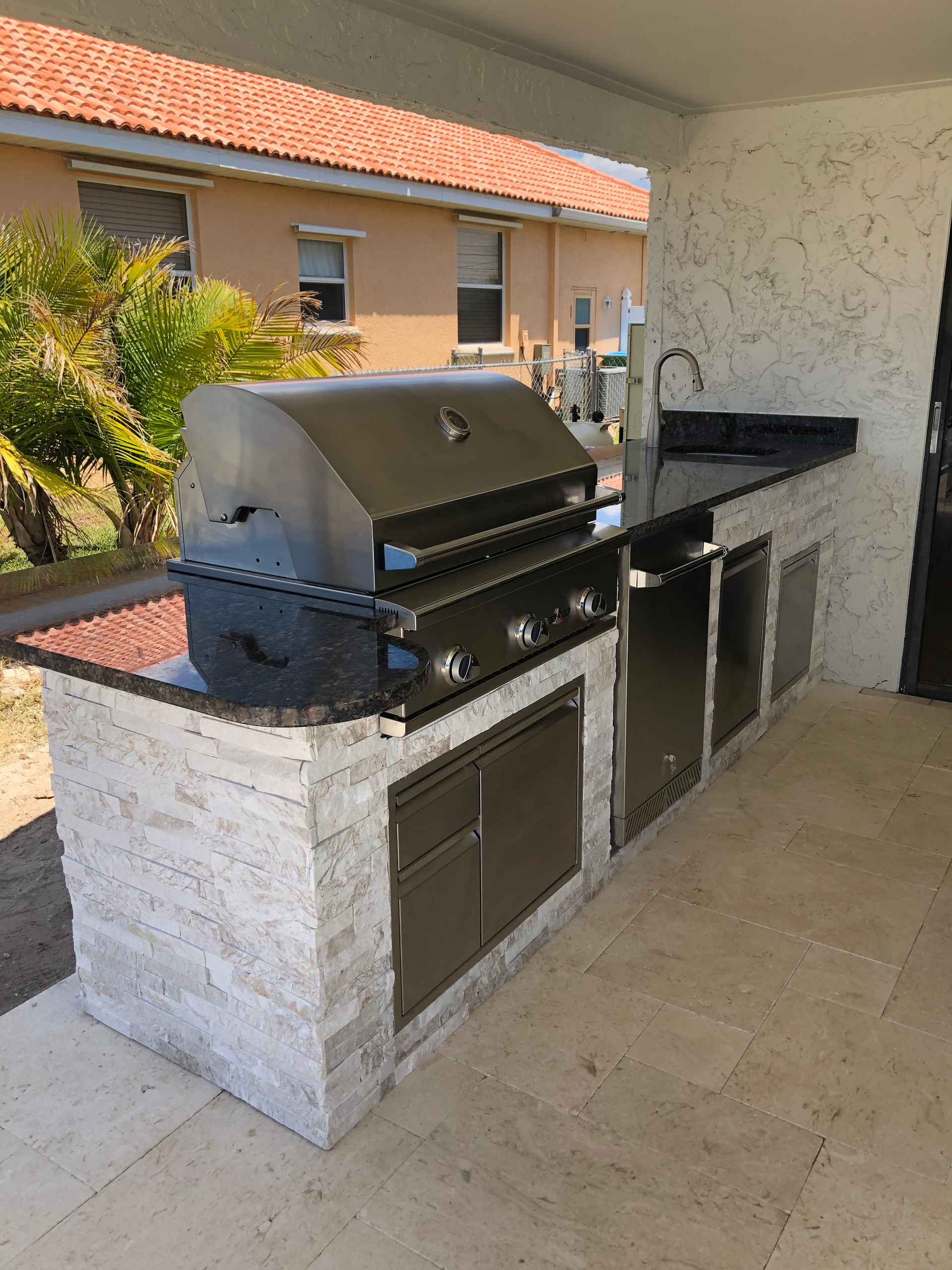 Outdoor kitchen with black grill, sink, cabinets, and a black countertop. Light brick facade and concrete patio.