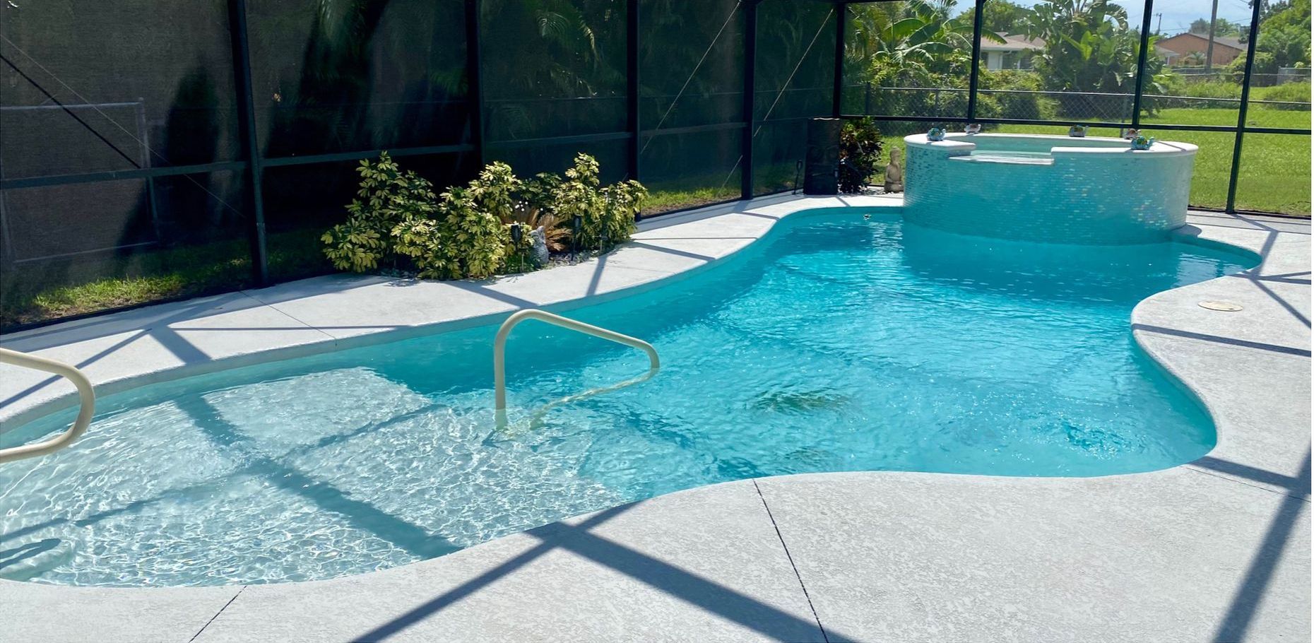A swimming pool with a built-in spa is surrounded by a light-colored patio enclosed by a screen.
