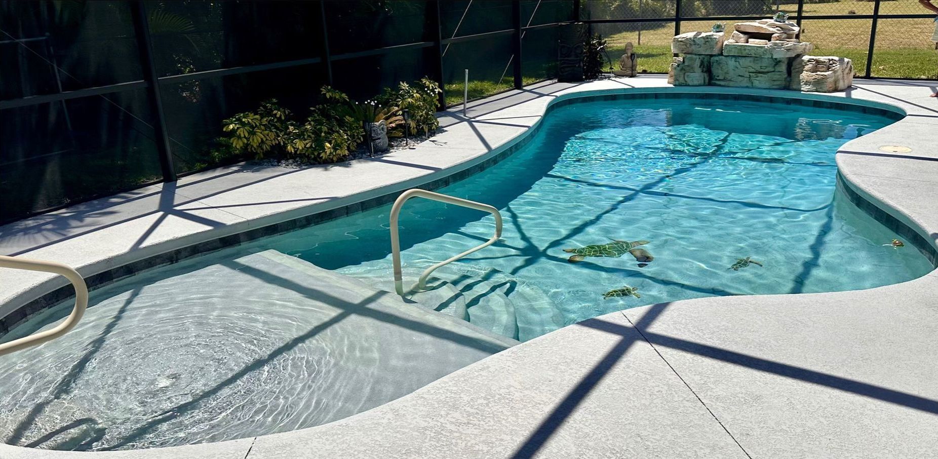 Pool with a stone waterfall feature, surrounded by concrete and a black fence. Blue water.