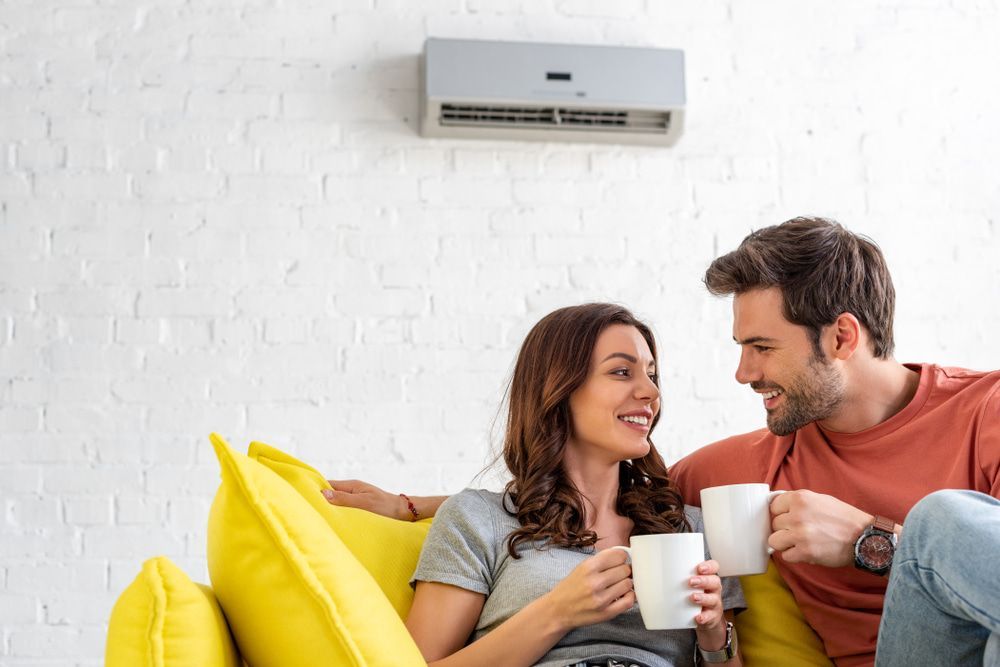 A Smiling Couple Relax on a Yellow Couch Under an Air Conditioner — Frosty Solutions Air Conditioning In Rylstone, NSW