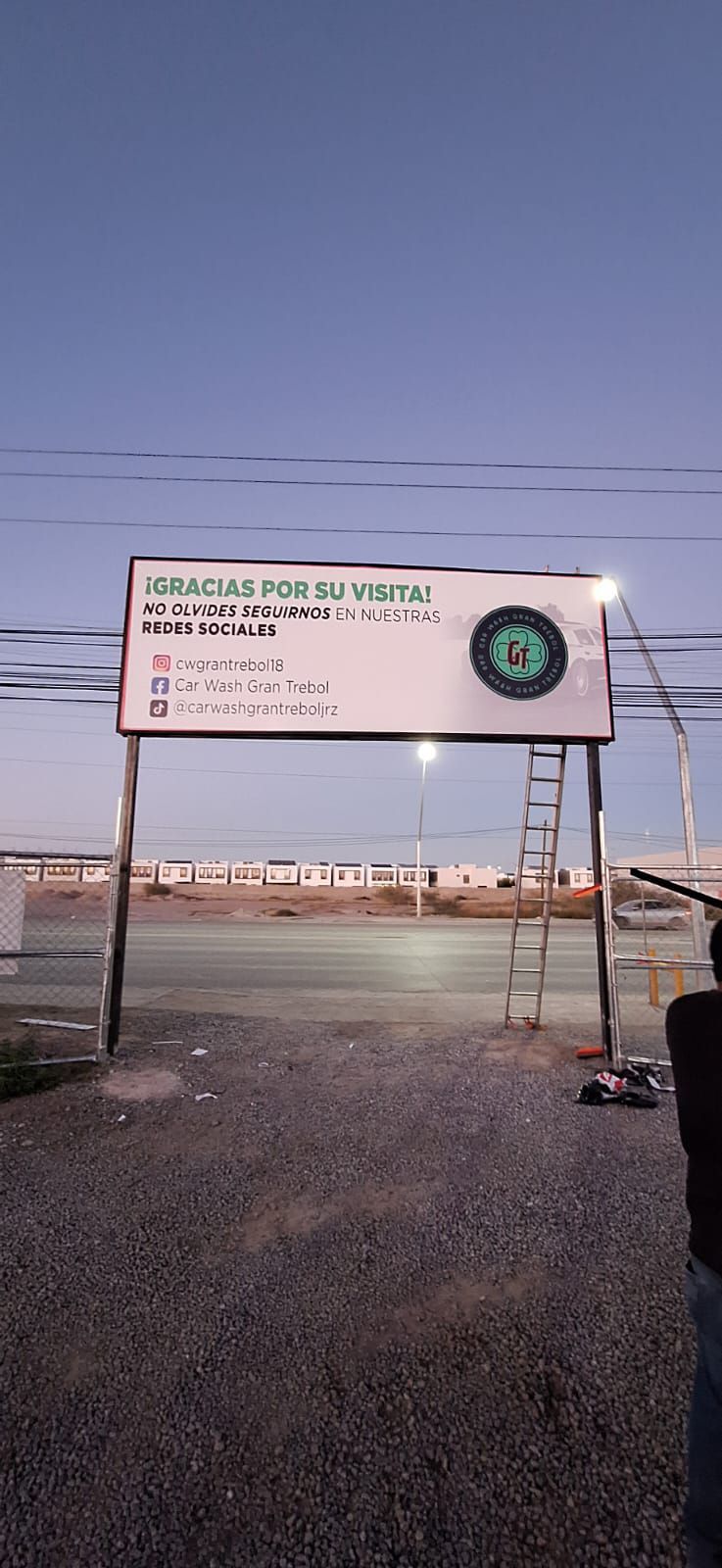 Letrero con texto y logotipo frente a un camino de grava y edificios bajo un cielo oscuro.