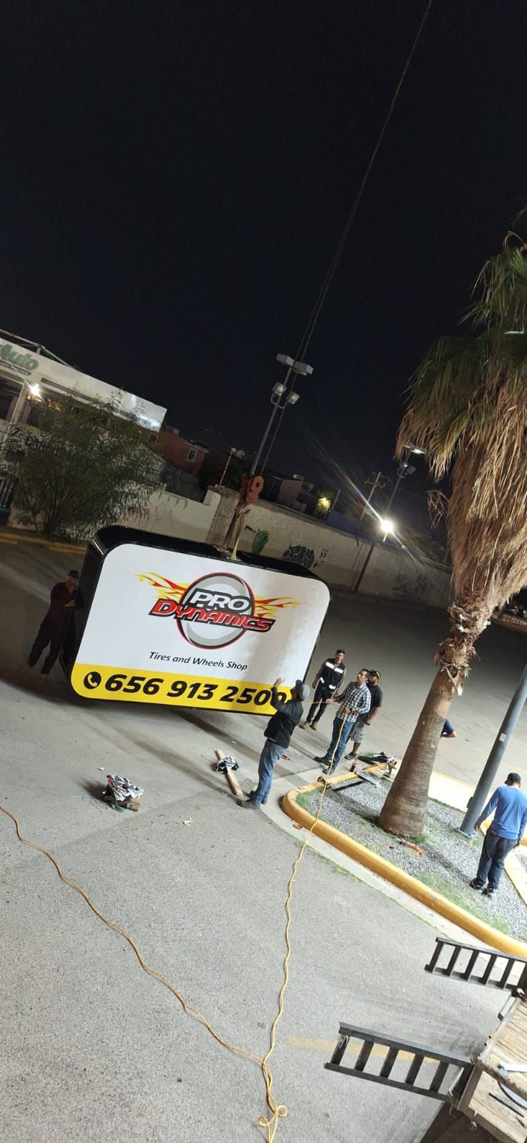 Escena nocturna: camión con un cartel, gente parada en una calle, cables eléctricos y un árbol.