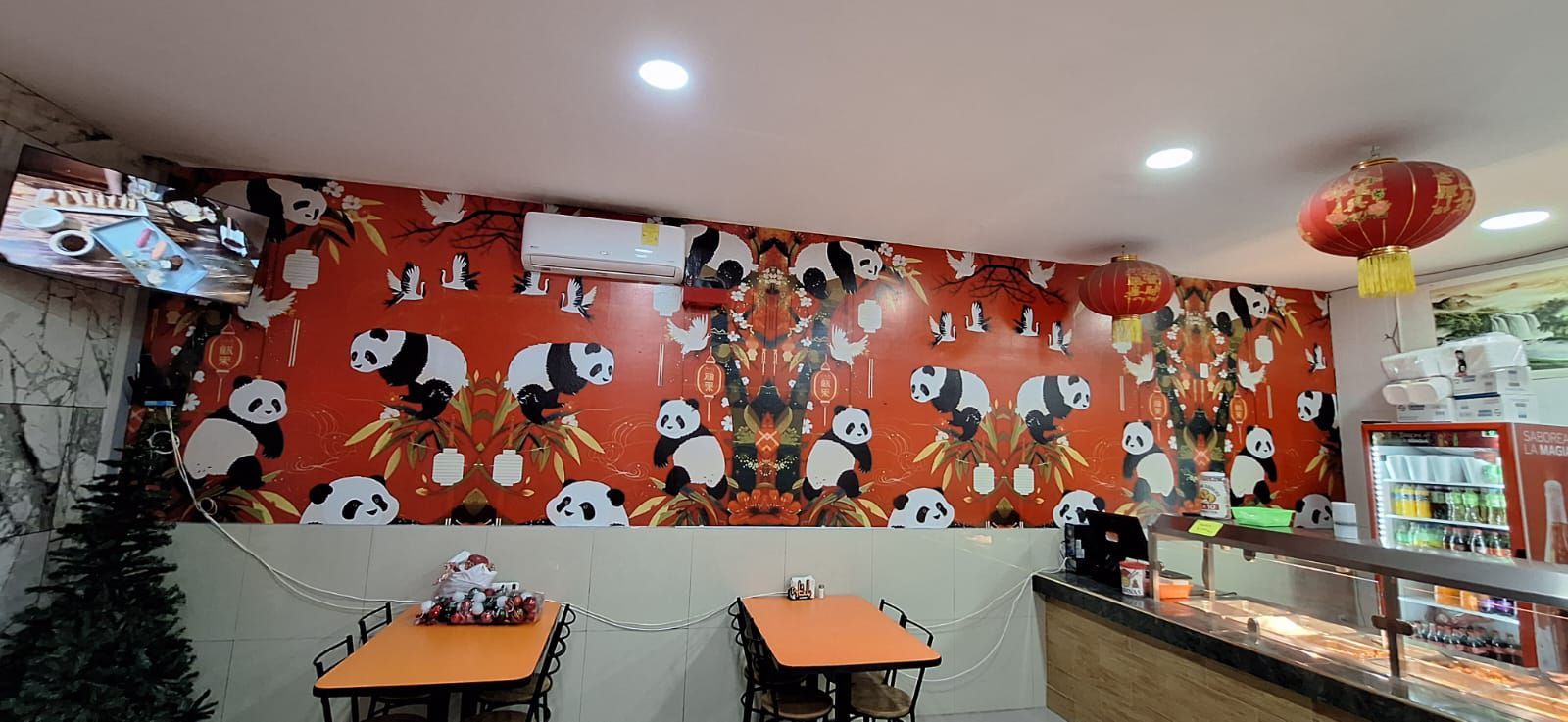 Interior de un restaurante con paredes y mesas con temática de pandas. Faroles rojos colgados y un refrigerador de bebidas visible.