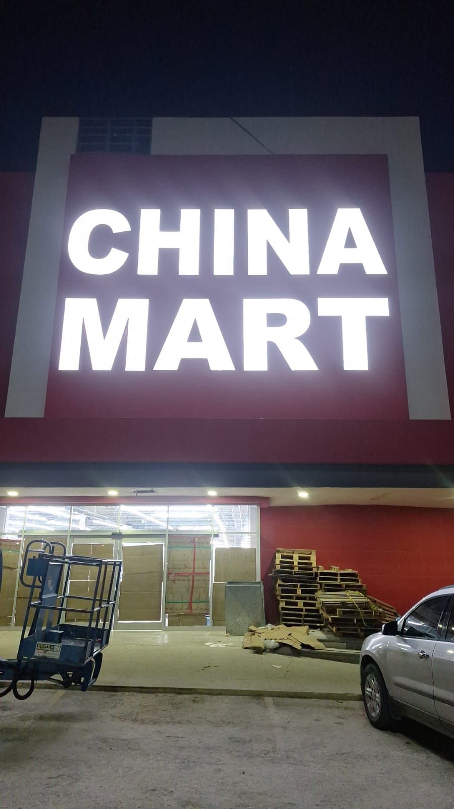 Entrada de la tienda China Mart, letras blancas iluminadas sobre fondo rojo. Cajas y un ascensor afuera.