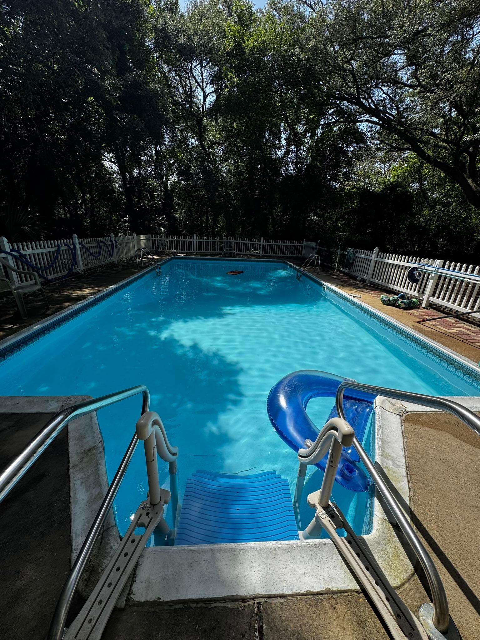 Outdoor swimming pool with blue water, steps, and a blue inflatable ring. Trees and a white fence surround the pool.