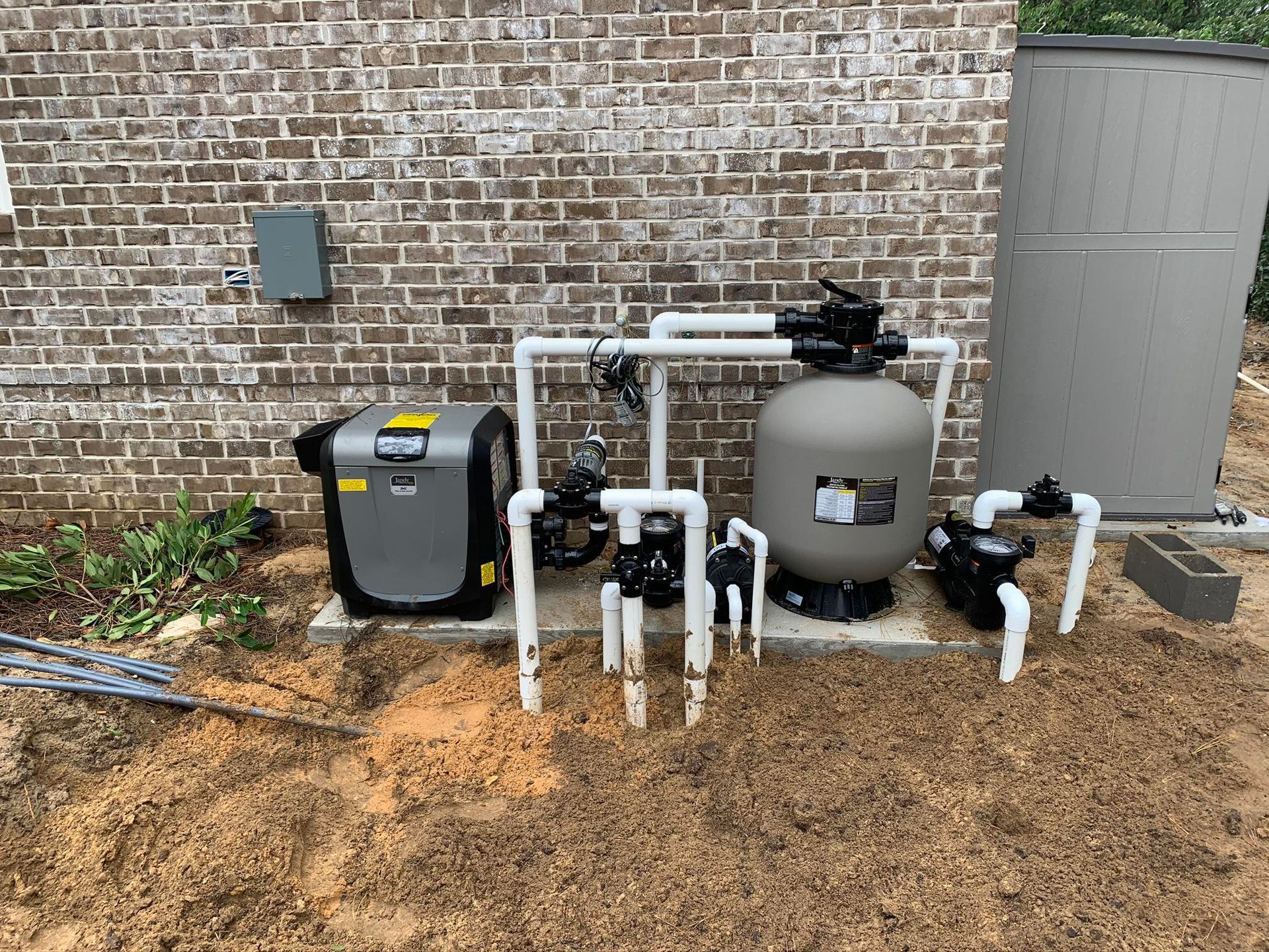 Pool equipment setup against a brick wall: a pump, filter, heater, and storage tank.