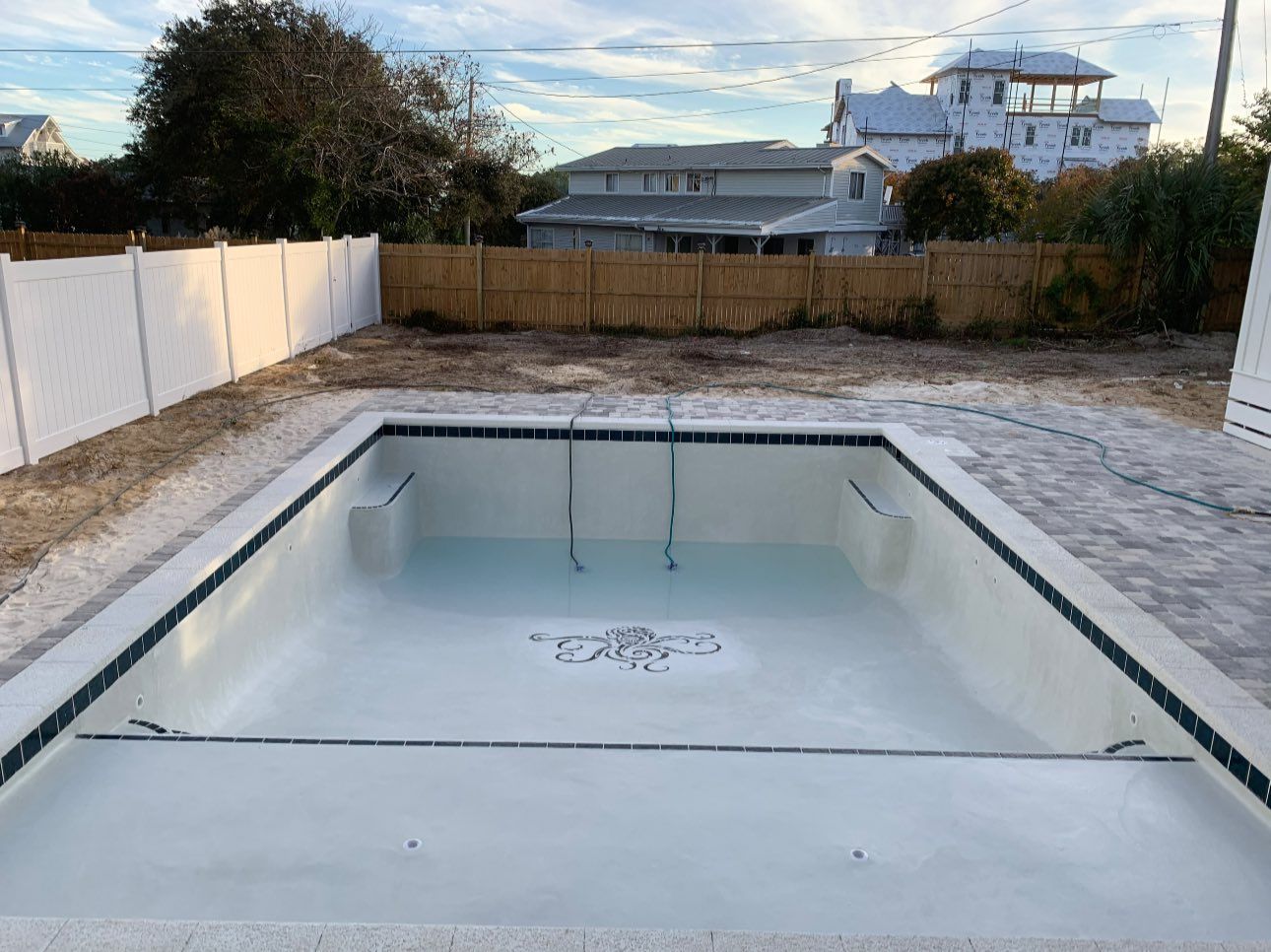 Empty rectangular swimming pool, light-colored, with a decorative tile emblem, surrounded by a patio and fences.