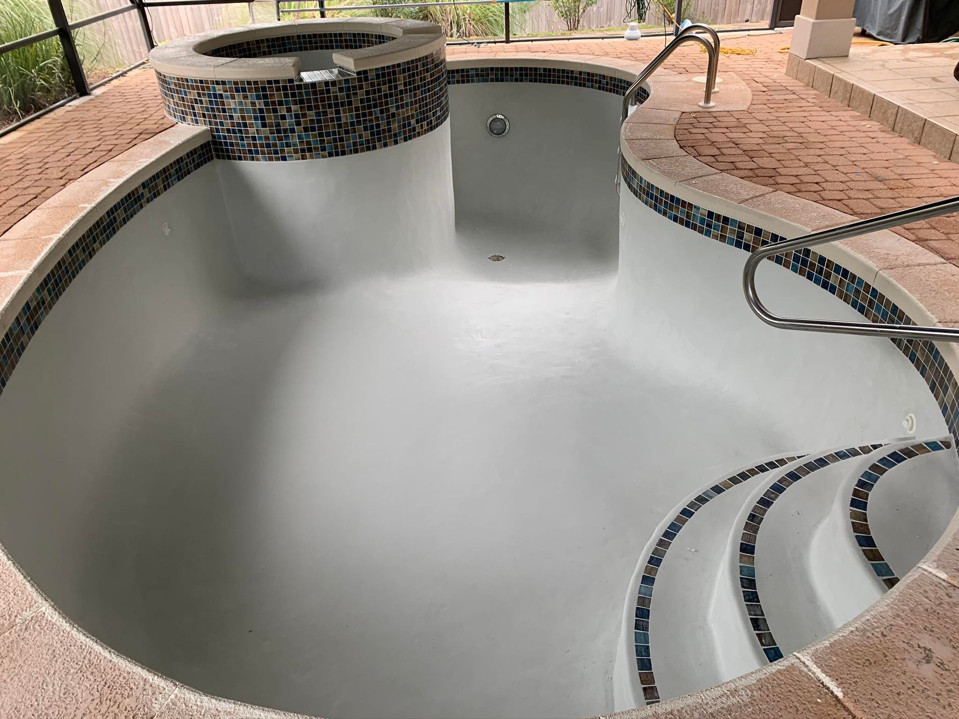 Empty, recently resurfaced pool with integrated spa, steps, and mosaic tile border.