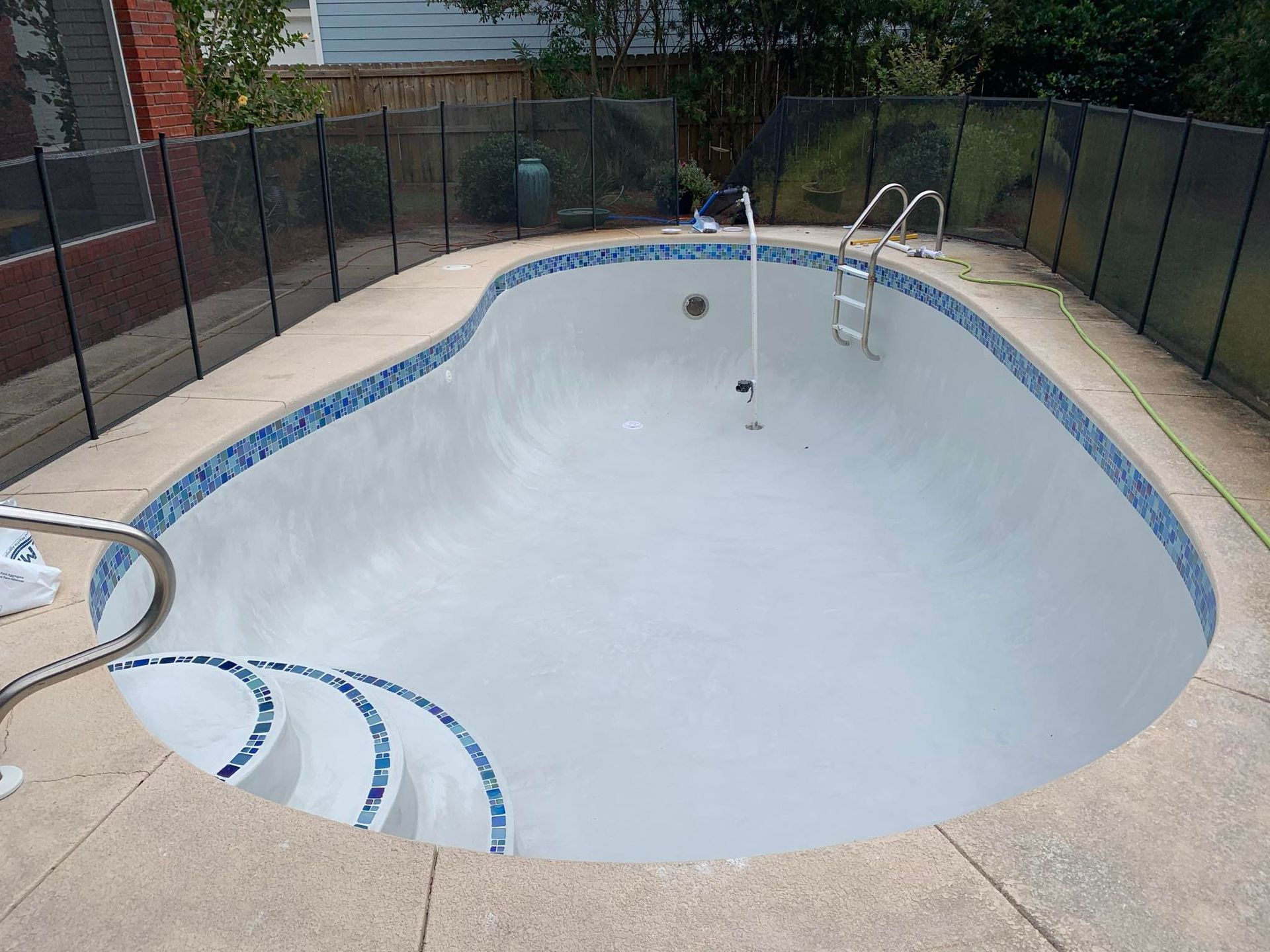 Empty, kidney-shaped swimming pool with blue tile trim, gray interior, and metal ladder. Black safety fence surrounds it.