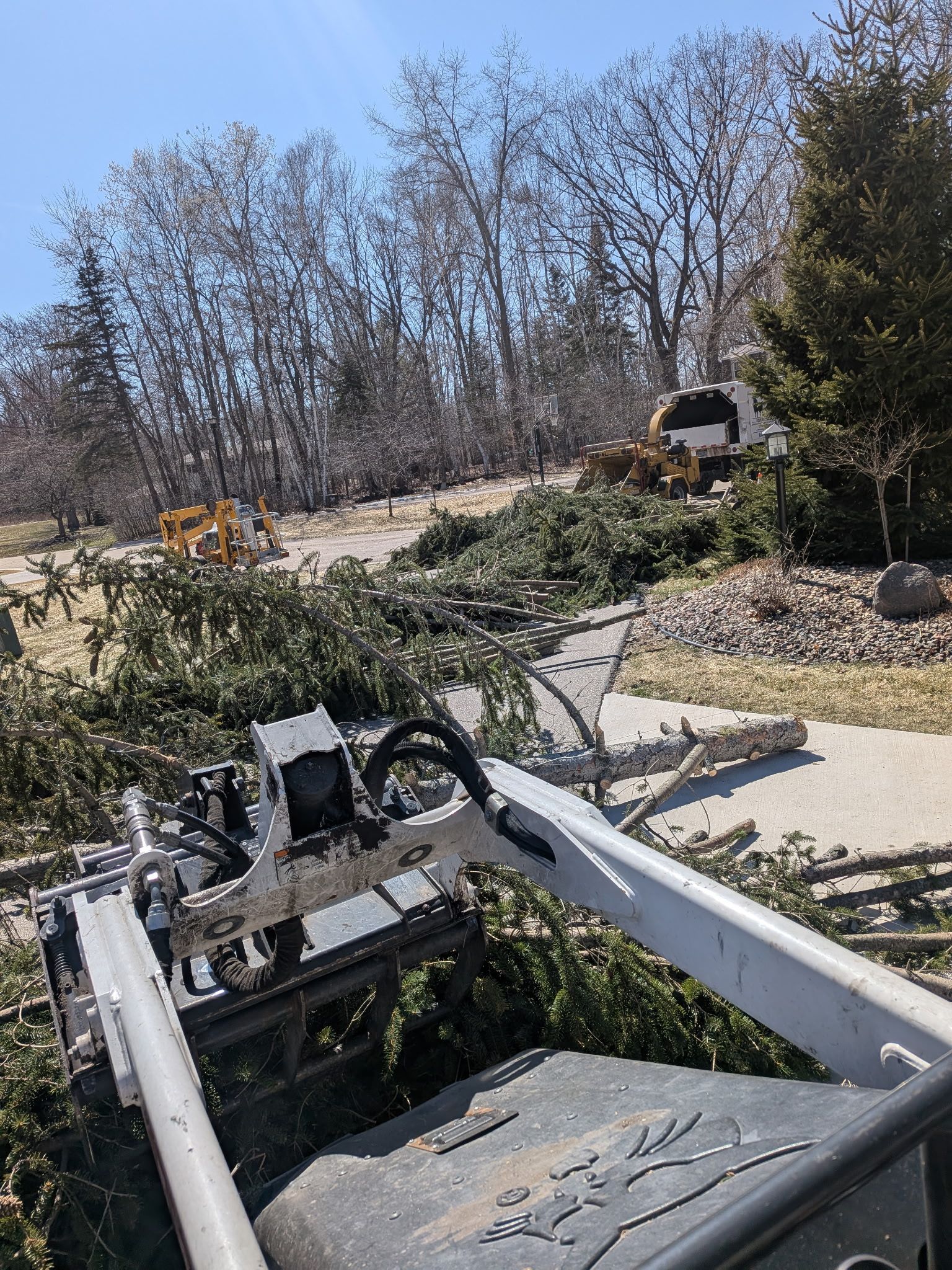 A skid steer loader attachment with a grapple is used to move cut tree branches on a sunny day.