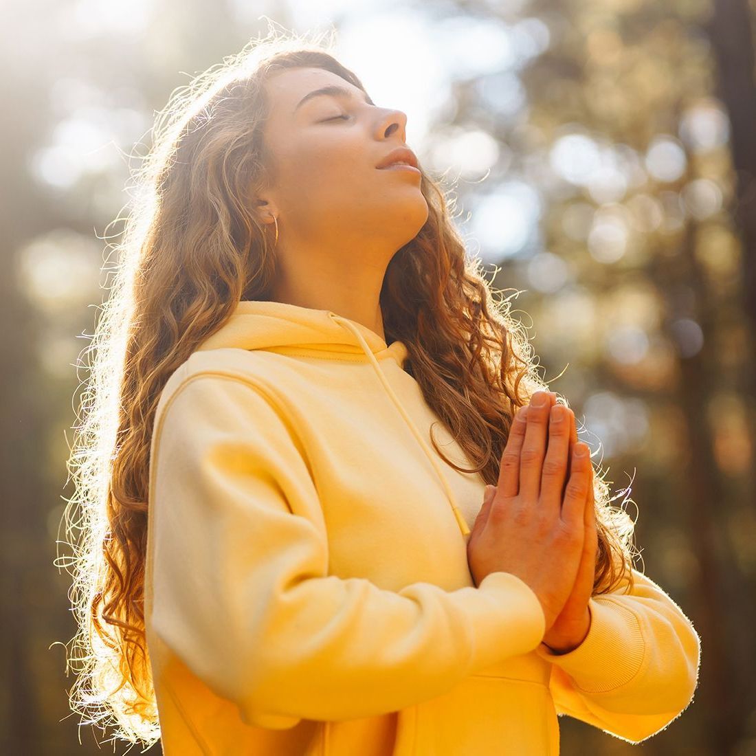 A woman in a yellow hoodie is praying with her eyes closed.