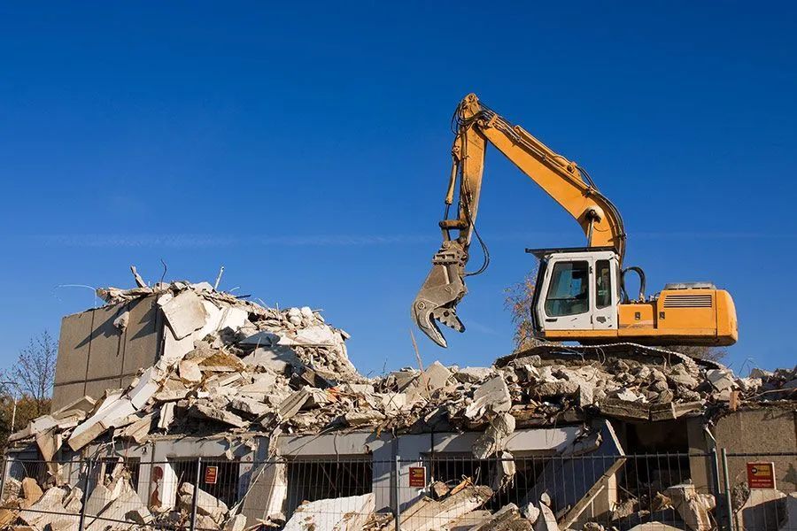 Yellow excavator demolishing a building under a clear blue sky. Debris scattered below.
