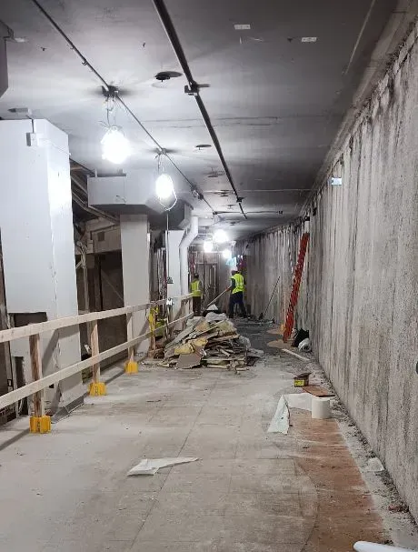 Construction workers clearing debris in a dimly lit, unfinished concrete corridor with overhead lighting and railings.