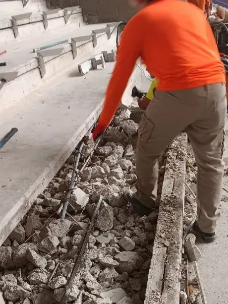 A person in an orange shirt works on exposed rebar in a rubble-filled trench at a construction site.