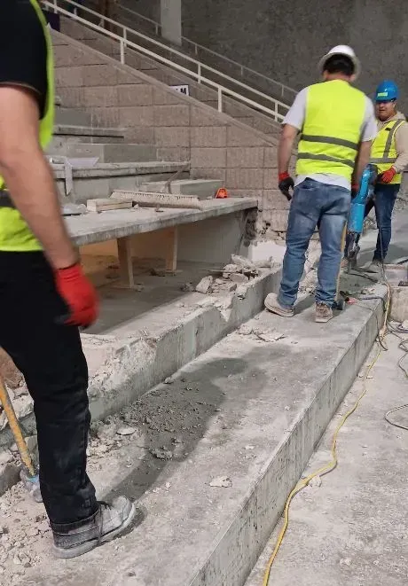Construction workers using a jackhammer to demolish concrete stadium steps.