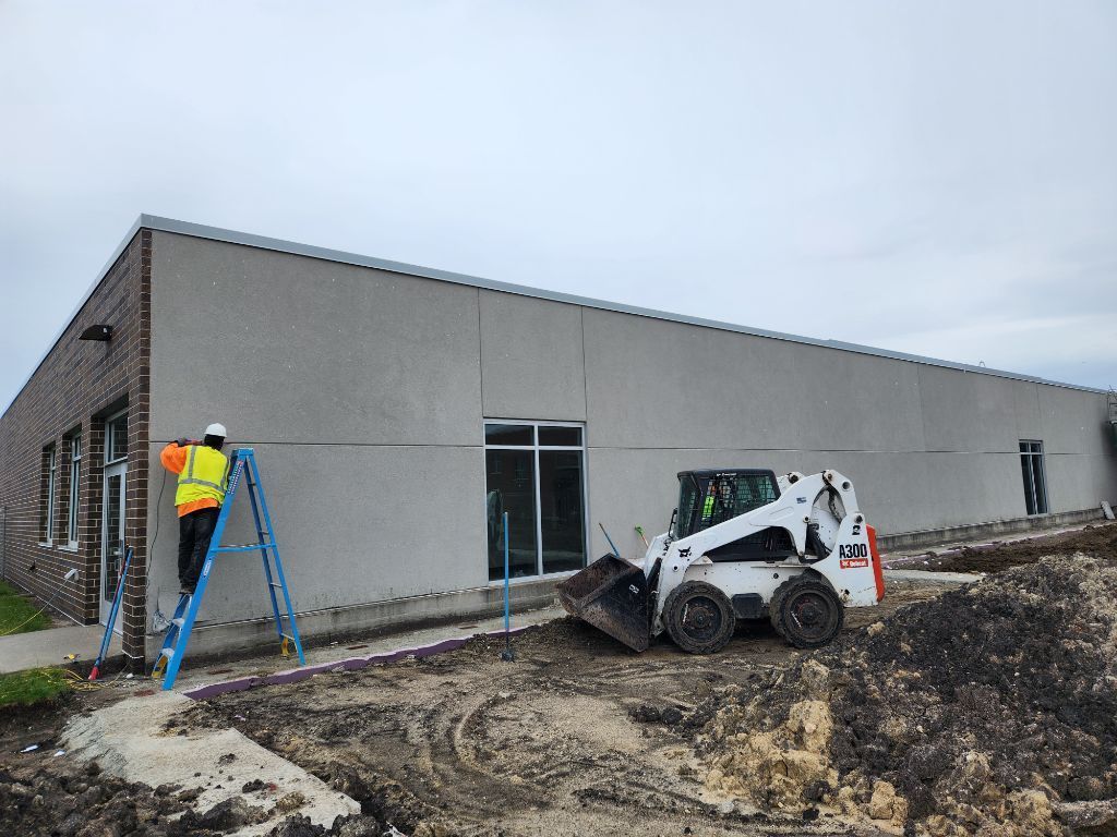 Workers use a skid steer and ladder outside a gray building under construction.