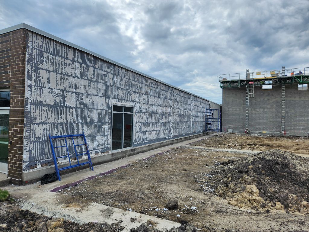 Construction site with a long concrete building, dirt foreground, and cloudy sky.