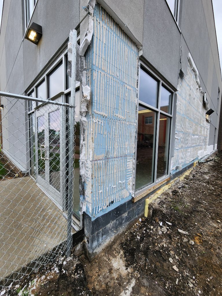 Damaged building corner with metal siding, glass doors, and a sloped concrete walkway.
