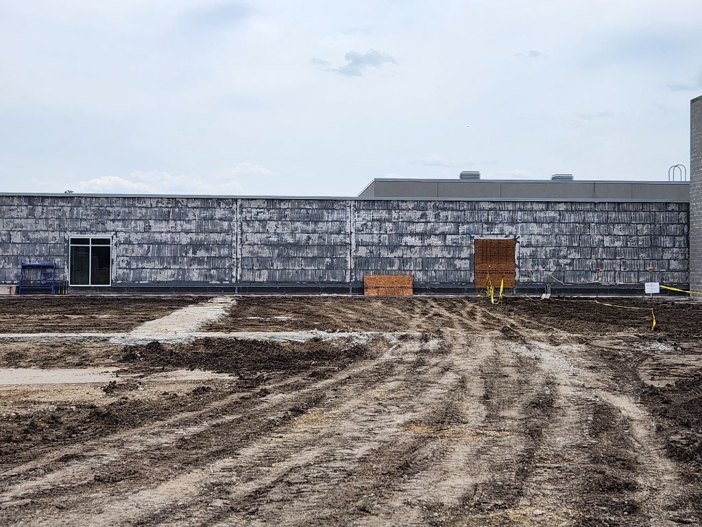 Gray industrial building on a muddy lot under a cloudy sky, with two orange dumpsters near the center.