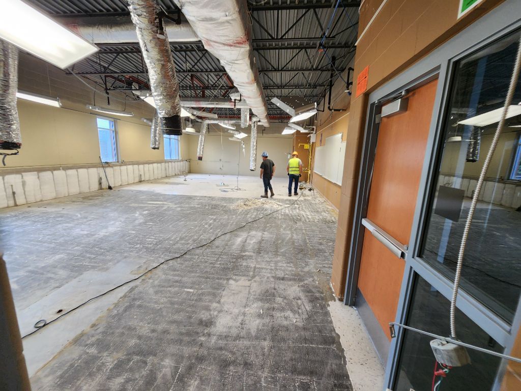 Indoor construction site with exposed ceiling ducts, unfinished floor, and two workers in the distance.