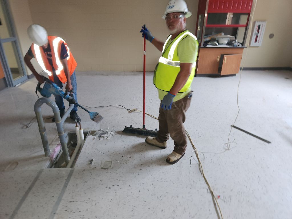 Two construction workers using jackhammers on a concrete floor indoors, one in a safety vest and hard hat.