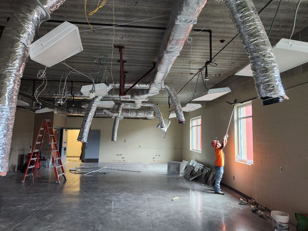 Interior of unfinished commercial space with exposed ducts, hanging lights, ladders, and construction materials