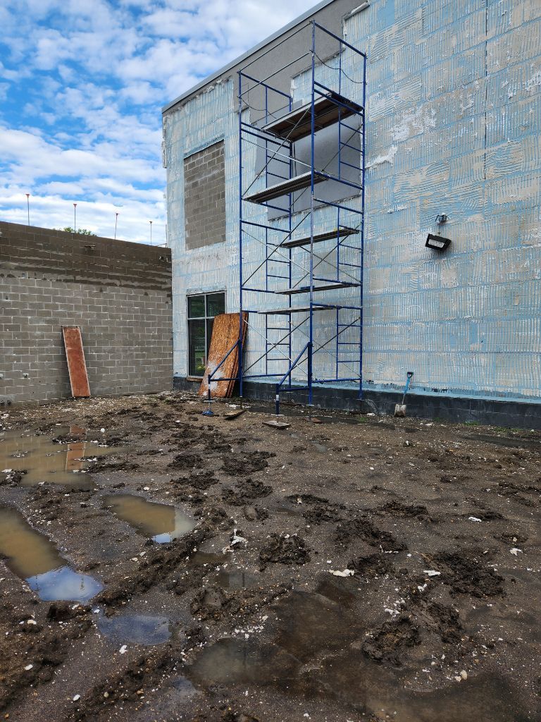 Scaffolding beside a light-blue building on a muddy construction site under a cloudy sky