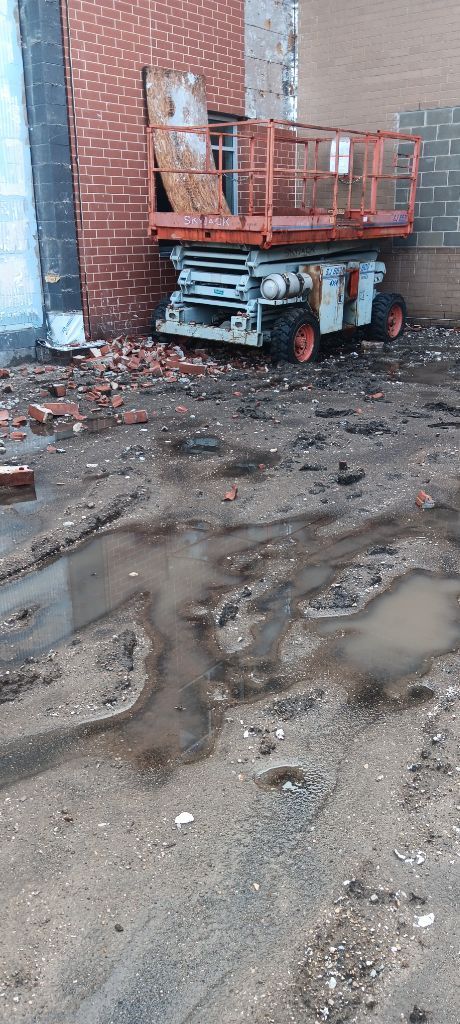Red scissor lift beside a brick wall on muddy, puddled ground outdoors