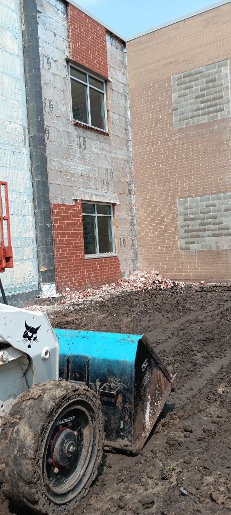 Blue skid steer beside a brick building, digging a muddy pile of dirt outdoors.